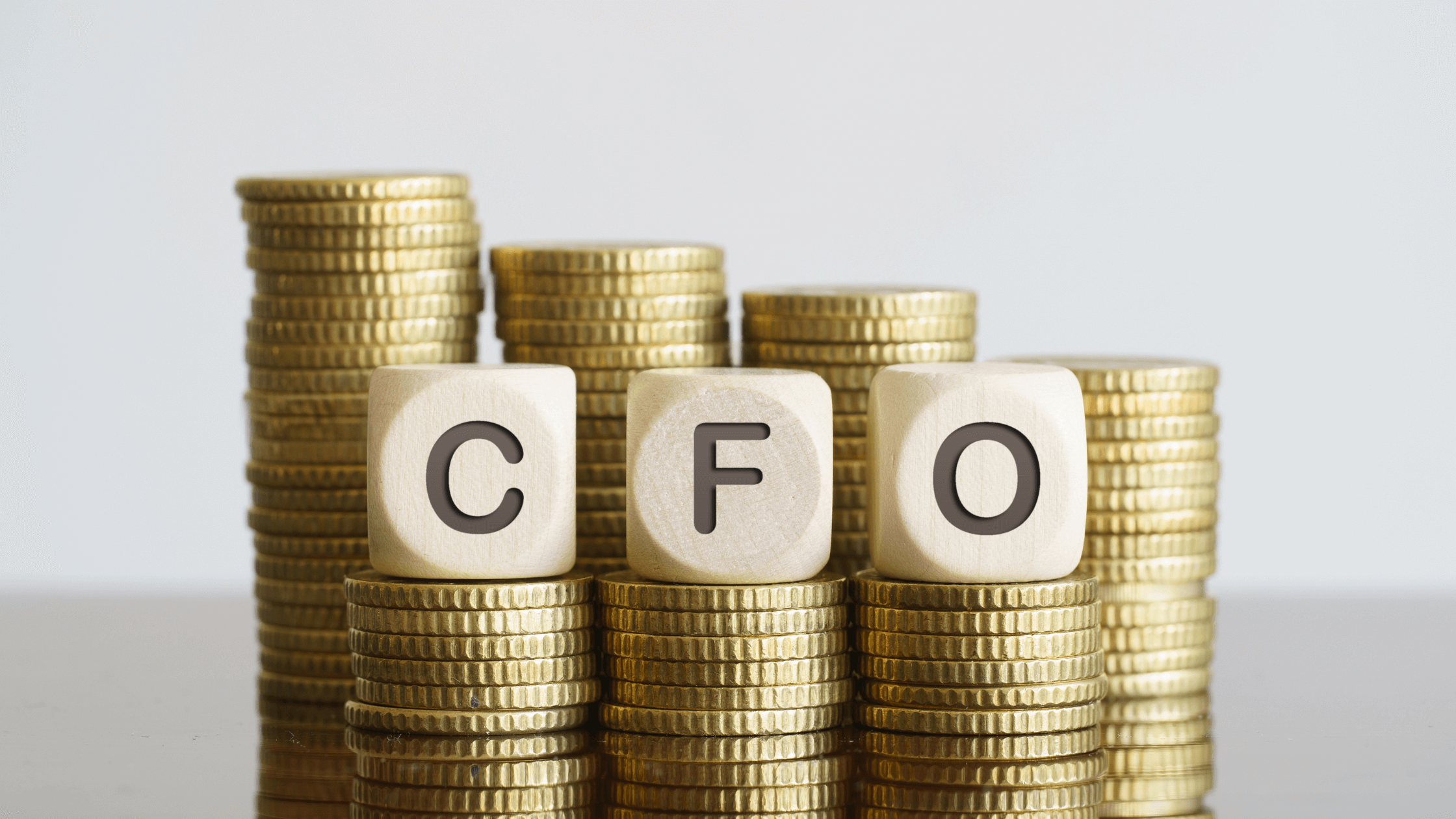 The letters C, F, and O are printed on wooden blocks stacked on top of a pile of gold coins, symbolizing the financial value and expertise a CFO brings to a business.