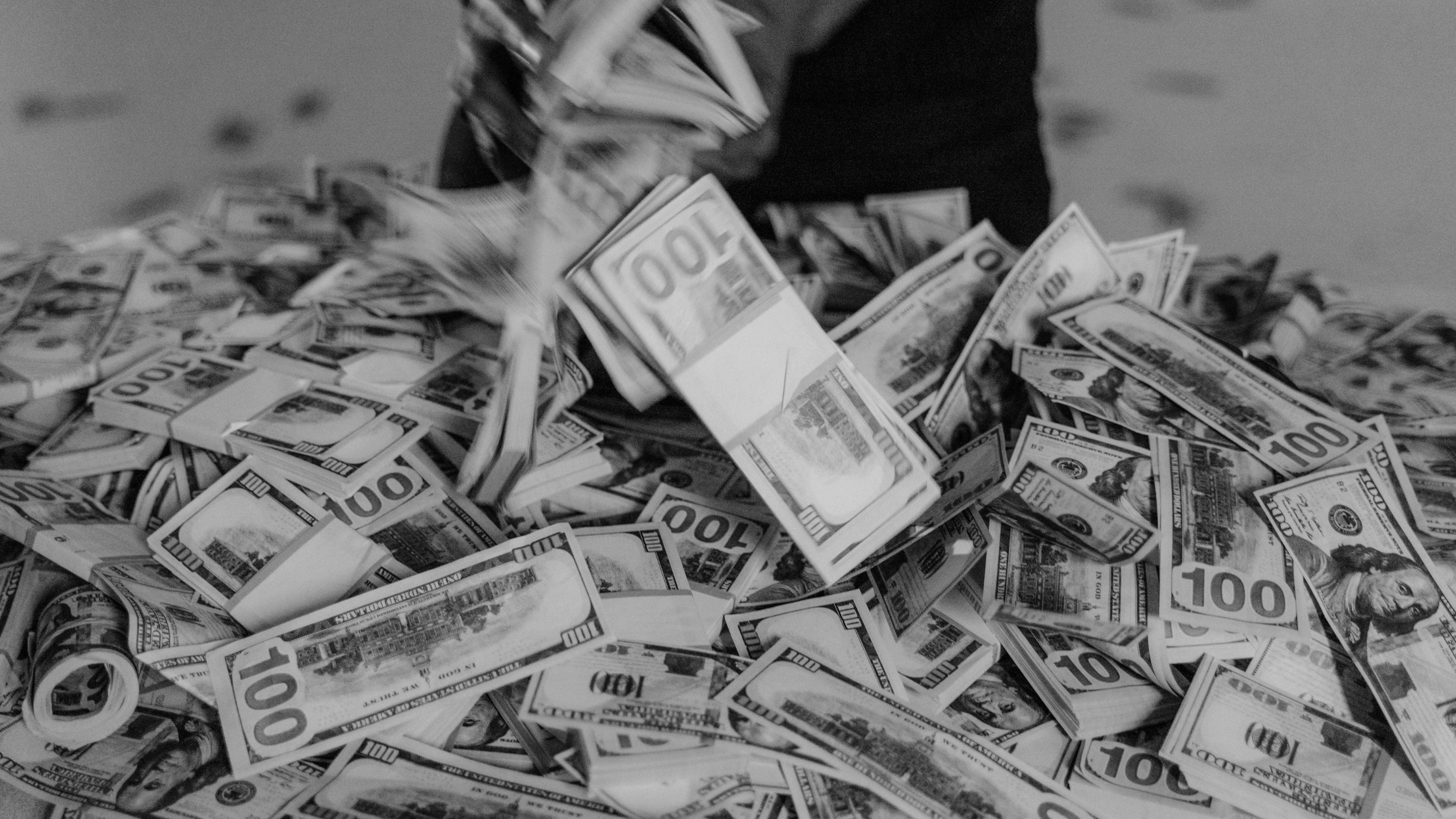 A pile of $100 bills in black and white, with hands tossing more cash onto the heap, representing the goal of high profitability and strong cash flow management for a small business.