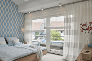 Cozy bedroom with white sheer drapery and patterned Roman shades on large balcony windows.