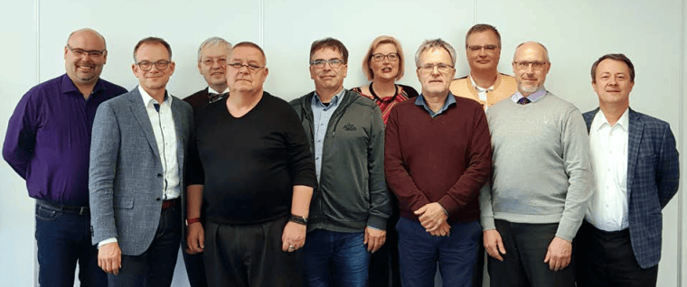 1. Mehrere Erwachsene stehen zusammen vor einer weißen Wand, lächeln und tragen formelle Kleidung, symbolisieren Teamarbeit bei Advent-Verlag.