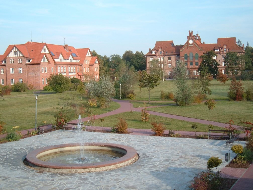 Historisches Gebäude mit rotem Ziegeldach in einem gepflegten Park mit Brunnen, Bänken und Gehwegen, ideal für Veranstaltungen und Erholung, verbunden mit dem Angebot von Advent-Verlag.