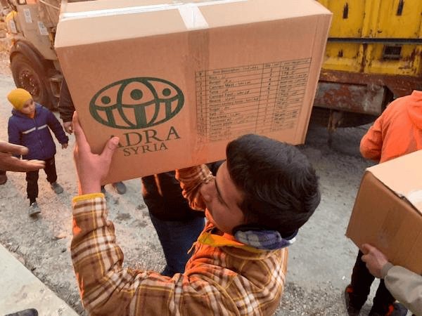 Große Karteikarte mit der Aufschrift "Adra Syrien" wird von einem Jungen beim humanitären Einsatz getragen, um die Versorgung in Krisengebieten zu unterstützen.