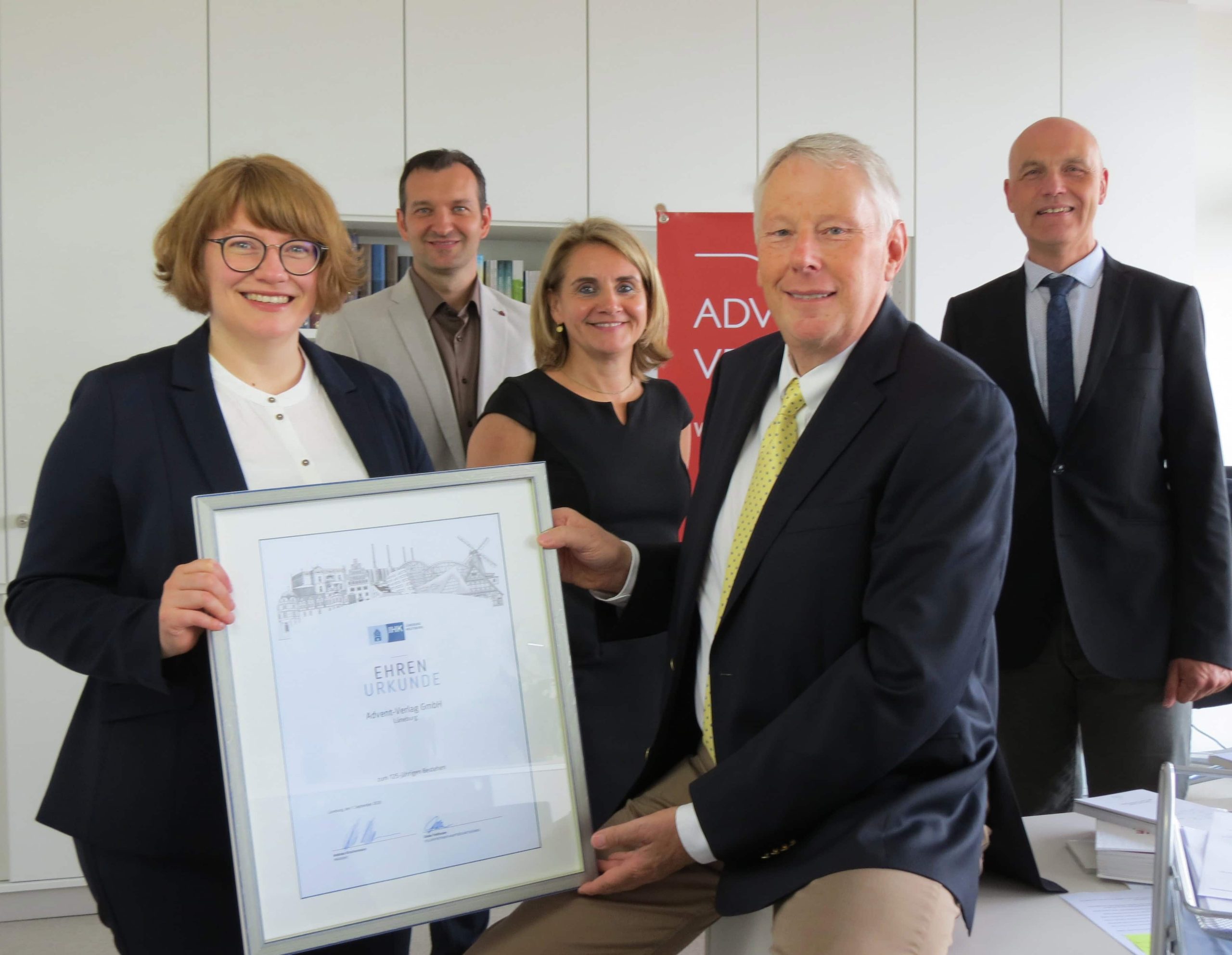 1. Gruppenfoto mit Mitarbeitern von Advent-Verlag bei Preisübergabe, Büro mit Büchern und Banner im Hintergrund, professionelle Atmosphäre.