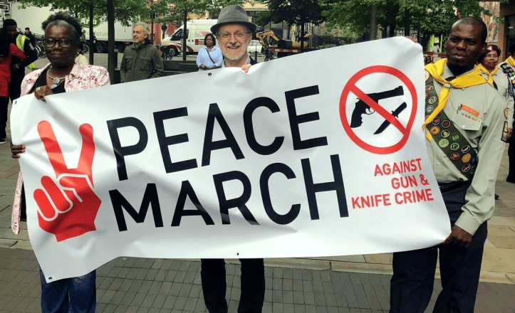 Friedensmarsch mit Protestbanner gegen Waffengewalt und Messerkriminalität, inklusive Friedenszeichen und Anti-Waffen-Logo, in einer urbanen Umgebung für Gewaltprävention und soziale Gerechtigkeit.
