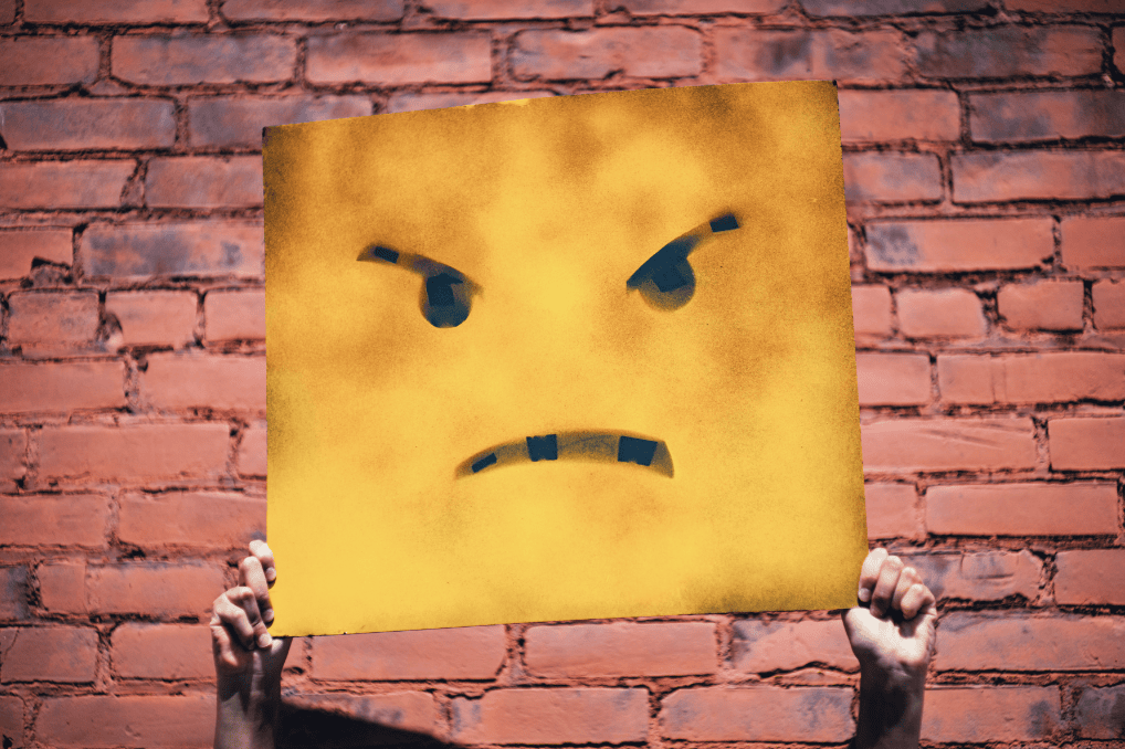 Wütendes Gesicht auf einem gelben Schild vor einer roten Ziegelmauer, symbolisiert Ärger und Frustration, passend für Konfliktlösungen oder emotionale Ausdrücke.