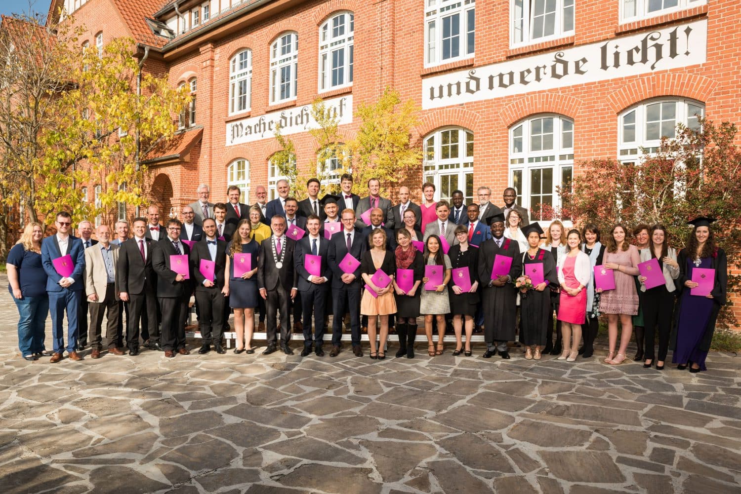 Erfolgsgeschwader mit Absolventen und Lehrkräften vor roten Backsteingebäude, Feierliche Abschlussfeier, Bildung, Studium, Abschluss, Universität, Erfolg, Gemeinschaft, Jubiläum, Unterricht.
