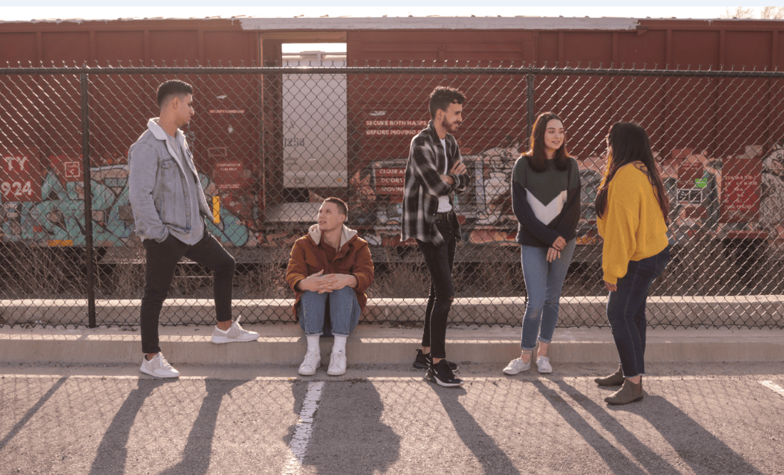 Jugendliche reden und lachen an einer Bushaltestelle, im Hintergrund ein Bahnwagon, in einem städtischen Umfeld, moderne urbane Atmosphäre, freundliche Gesellschaft, Street Style Mode, junge Menschen, Freizeit, urbaner Lifestyle, Jugendliche Treffpunkt, Zusammenhalt.