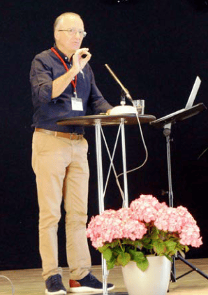 1. Sprecher bei einer Präsentation auf einer Bühne, hinter einem Pult mit Laptop, umgeben von pinken Hortensien, bei einer Veranstaltung von Advent-Verlag.