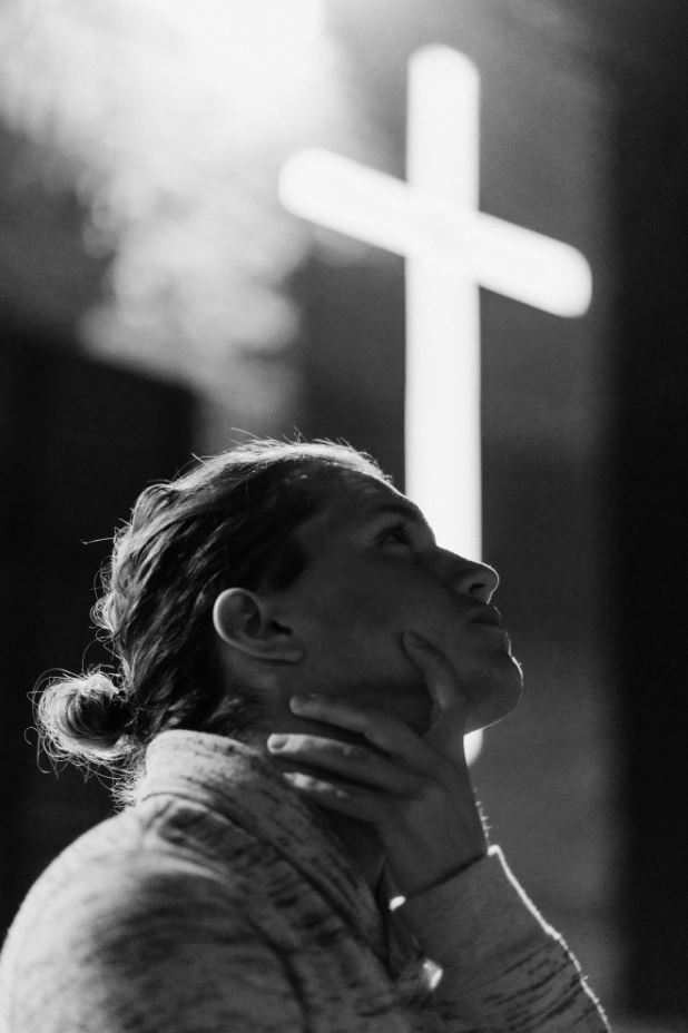 Gebet einer jungen Frau vor einem hell leuchtenden Kreuz in einer Kirche, Schwarz-Weiß-Aufnahme.