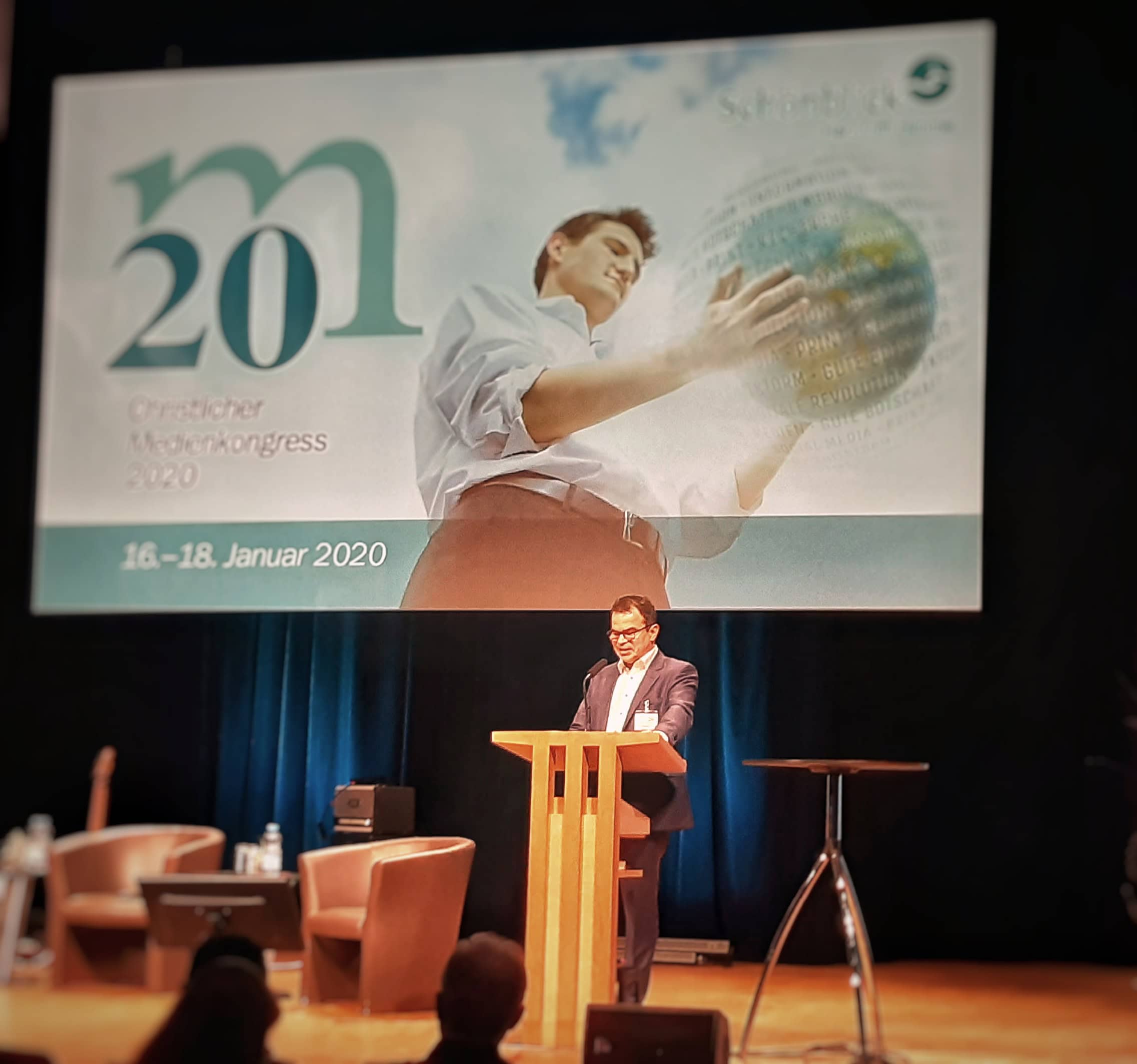 1. Hochwertiges Bild eines Redners auf dem Medizin Kongress 2020, mit großem Präsentationsbild und Sitzmöbeln, Fokus auf medizinischer Fortbildung.
