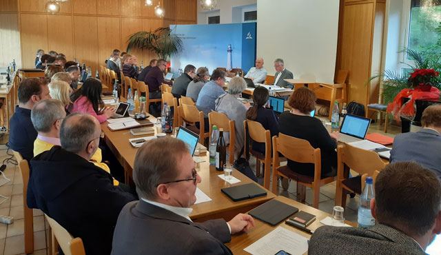 Innovatives Verlagswesen bei Advent-Verlag: Conference mit Teilnehmern, die an Laptops und Tablets arbeiten, in einem Raum mit natürlichen Licht und Meeresbilder im Hintergrund, Fokus auf geistiges und kreatives Arbeiten.