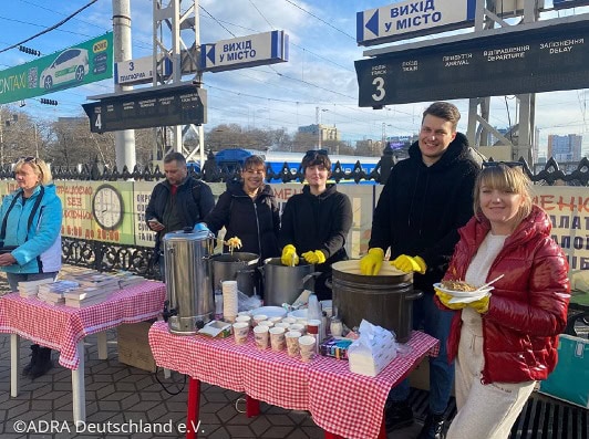 Frühstücksstand am Bahnhof mit Kuchen und warmen Getränken, engagierte Helfer in Gelbhandschuhen, freundliche Veranstaltung, ideal für Gemeinschaftsaktionen oder Spendenaktionen entlang der Bahnlinie.