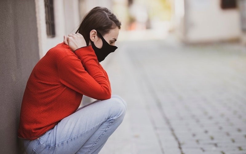 1. Junge Frau mit schwarzer Maske sitzt traurig auf der Straße, urbaner Hintergrund, Herbst, nachdenklich, emotional, Pandemie, Sorgen, Isolation, Ängste.