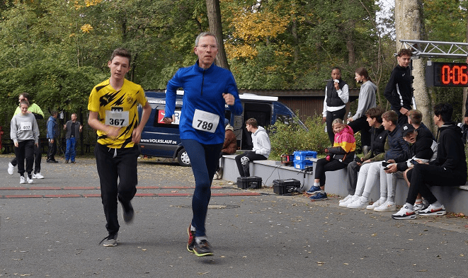 1. Läufer beim Straßenlauf im Park, Ergebnisanzeige im Hintergrund, herbstliche Szene, gehostet von Advent-Verlag.