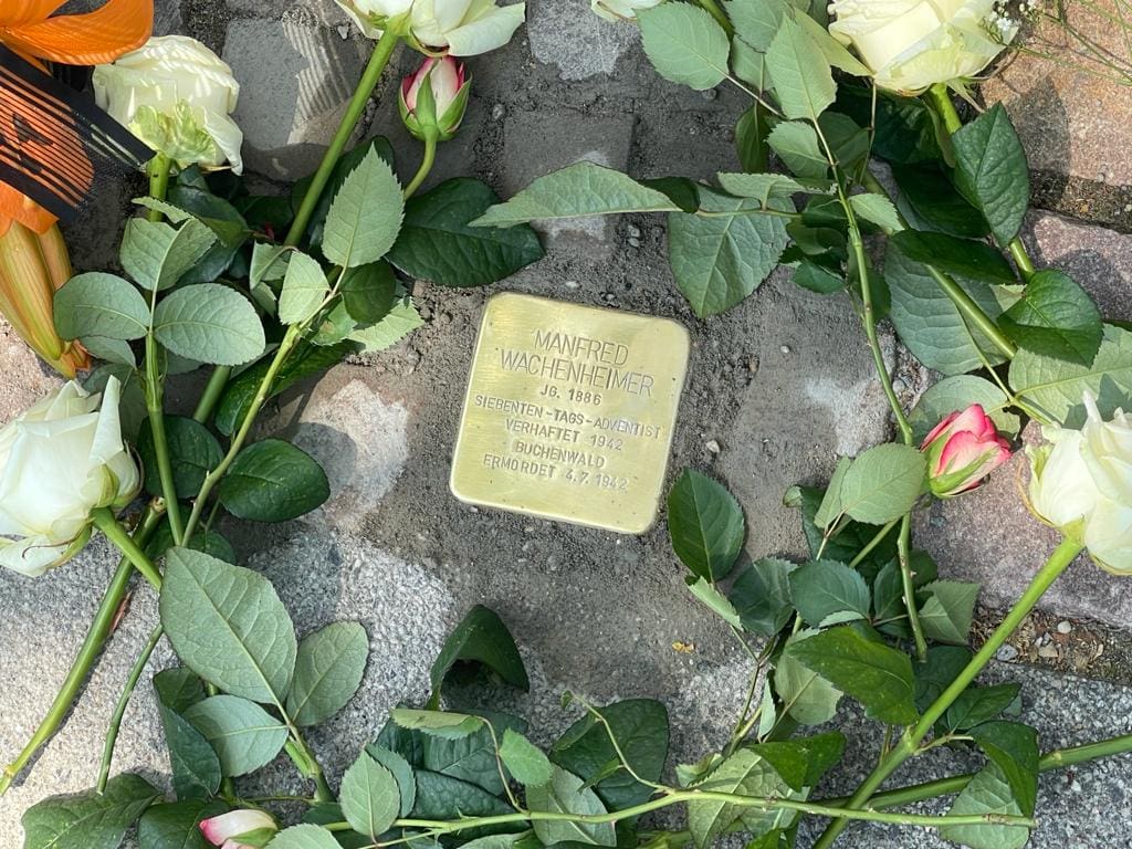 Gedenkstein für Manfred Wachenheimer, umrahmt von weißen Rosen, auf dem Straßenpflaster, symbolisiert Erinnerung und Ehrung eines Verstorbenen.