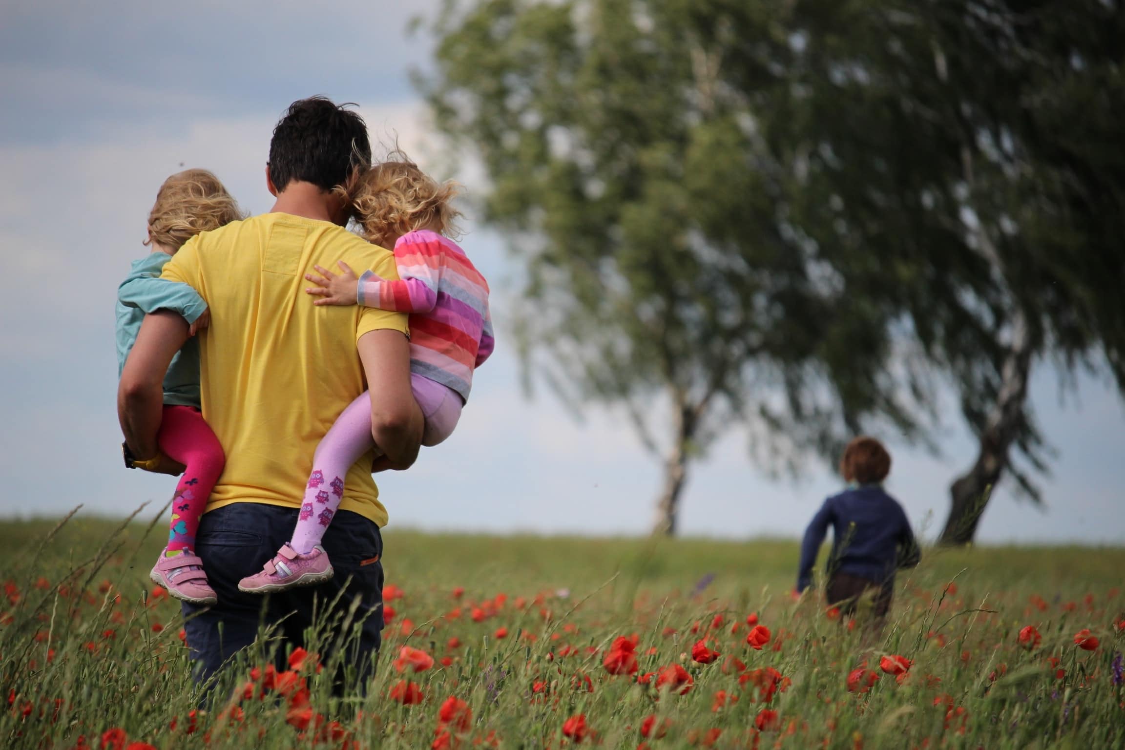 Fröhliche Familie beim Spaziergang in der Natur, Kinder auf den Schultern der Eltern, umgeben von blühenden Wiesen und Bäumen, entspannte Freude im Grünen.