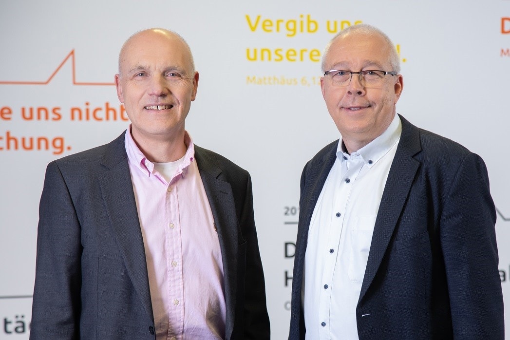 Menschen bei einer Veranstaltung, zweisitzige Portraits, professionelle Kleidung, Hintergrund mit Texten zu Glauben und Vertrauen, formelle und inspirierende Atmosphäre, vermutlich religiöse oder christliche Thematik, Konferenz oder Seminar, Mixtur aus Natur- und Bürowelten, freundliche Gesichter, Expertengespräch, Gemeinschaftsbildung.
