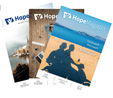 Drei HopeMagazin-Ausgaben des Advent-Verlags mit inspirierenden Titeln wie "Heimweh", "Fernweh" und "Sehnsucht" vor einem Holztisch, symbolisieren Hoffnung und Glauben.