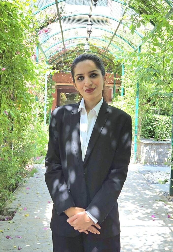 An image of Advocate Sonali Sardana standing confidently outdoors in a lush, green garden with a colorful arched canopy overhead, dressed in a professional black suit and white shirt.