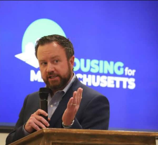 Man in a suit speaks into a microphone at a wooden podium, with a blue backdrop reading 'Housing for Massachusetts' behind him.