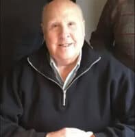 Smiling older man with short hair wearing a black zip-front pullover, seated indoors at a table.