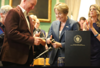 Woman in a blue blazer hands a ceremonial award to a man in a brown jacket at a formal ceremony, with a seal-emblazoned podium nearby.