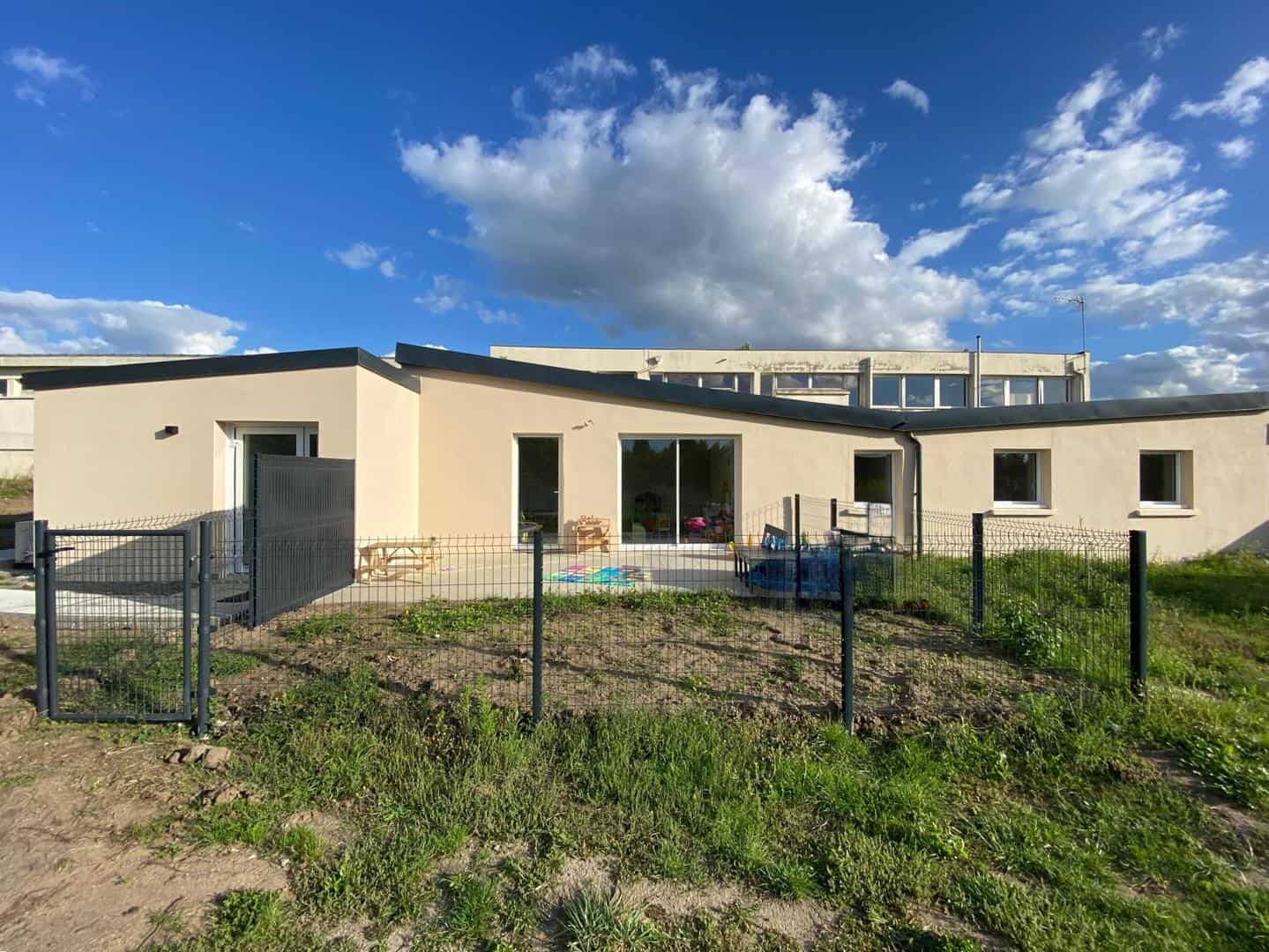 Photo de la façade d’un bâtiment récemment rénové, avec un crépi beige uniforme et une toiture en pente moderne. Un jardin clôturé entoure l’espace extérieur, et de grandes baies vitrées permettent une luminosité optimale. Le ciel bleu et dégagé met en valeur les finitions impeccables de la structure