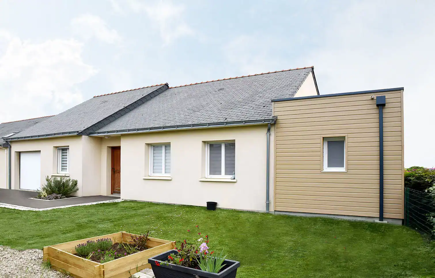 Une extension moderne et harmonieuse qui s’intègre parfaitement au style de la maison existante. Le bardage beige clair contraste élégamment avec la façade principale, créant un effet contemporain. Les finitions soignées, telles que les gouttières et les encadrements, renforcent l’aspect épuré. L’espace extérieur est optimisé avec une pelouse bien entretenue et un carré potager en bois, ajoutant une touche naturelle et fonctionnelle. Une réalisation esthétique qui apporte un nouvel espace de vie tout en valorisant l’architecture d’origine.