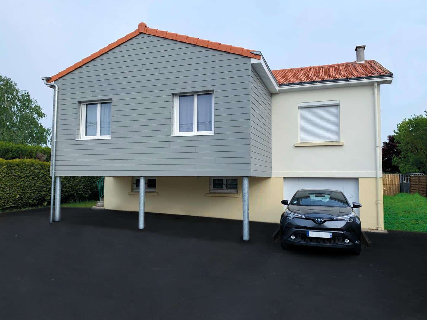 Maison avec une extension en bardage gris, voiture stationnée devant, et sous-sol sur pilotis.