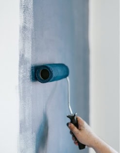 Close-up of a person using a paint roller to apply light blue paint on a wall.
