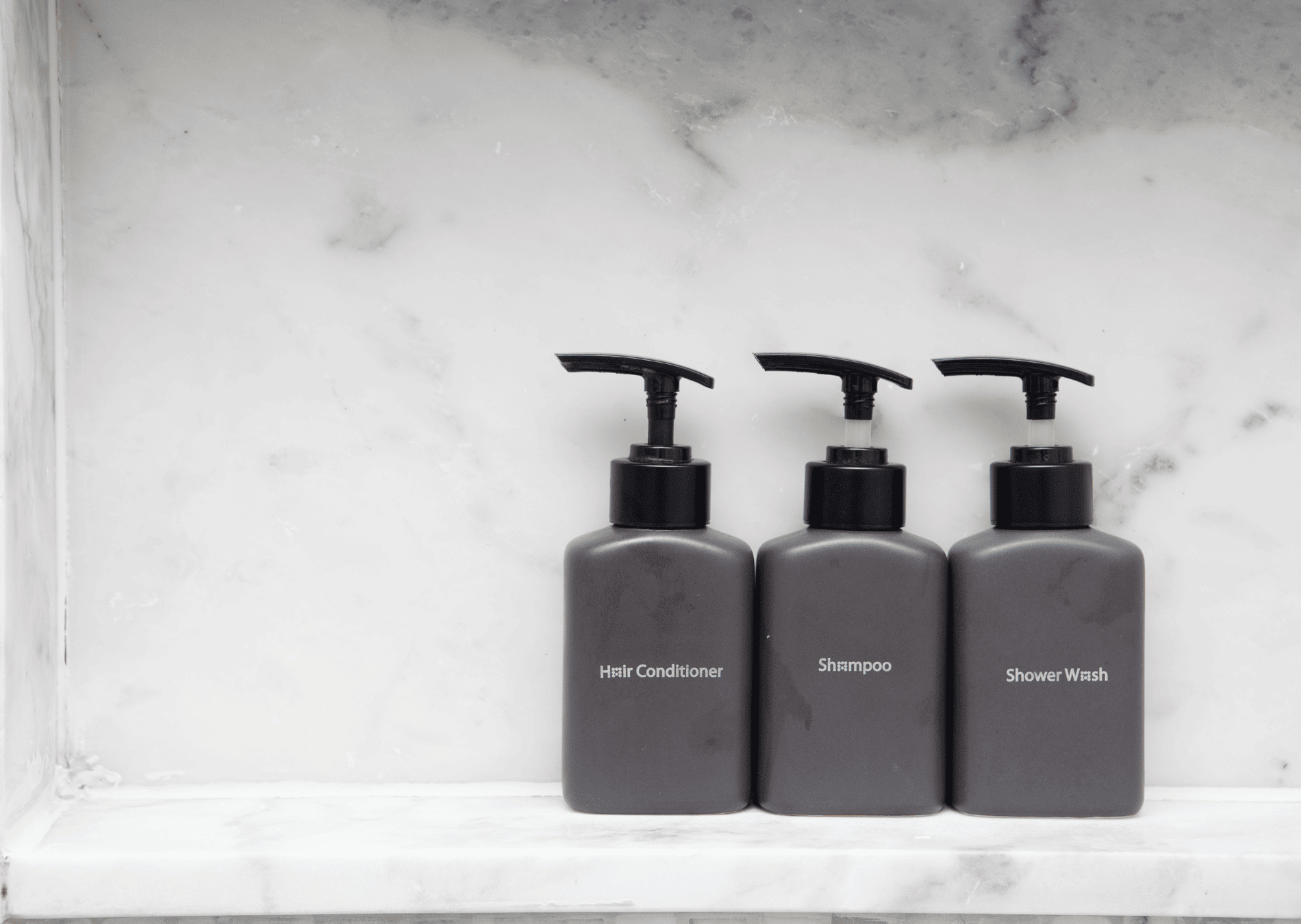 Drie zwarte hotelzeepdispensers met haarconditioner, shampoo en douchewash op een marmeren plank.