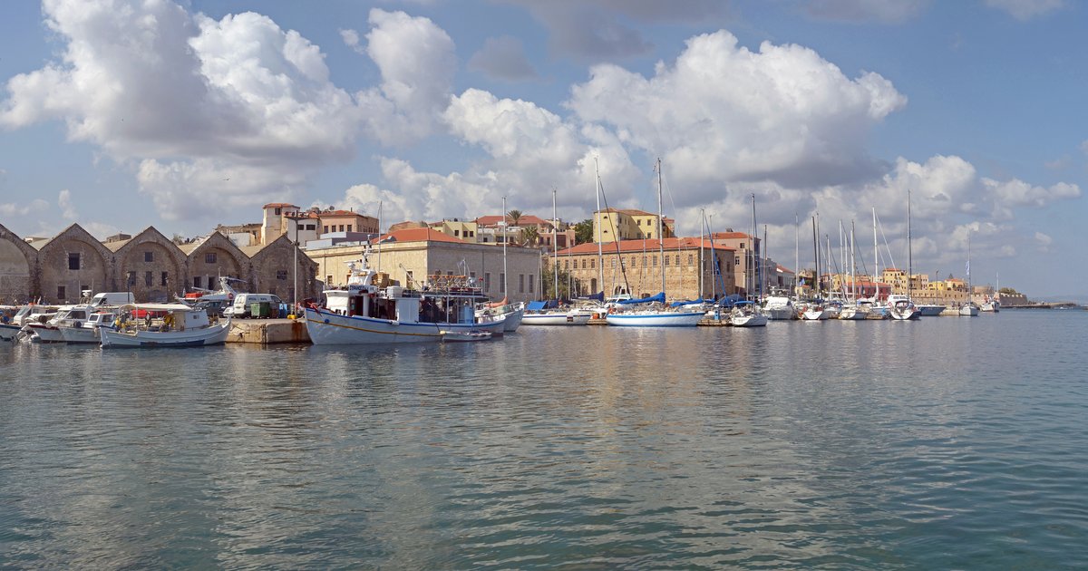 Τα ενετικά νεώρια και τα σκάφη στο παλιό λιμάνι των Χανίων κάτω από λευκά σύννεφα.