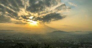Ο ήλιος ανατέλλει πίσω από σύννεφα πάνω από λόφους και μια πόλη στην ομίχλη.