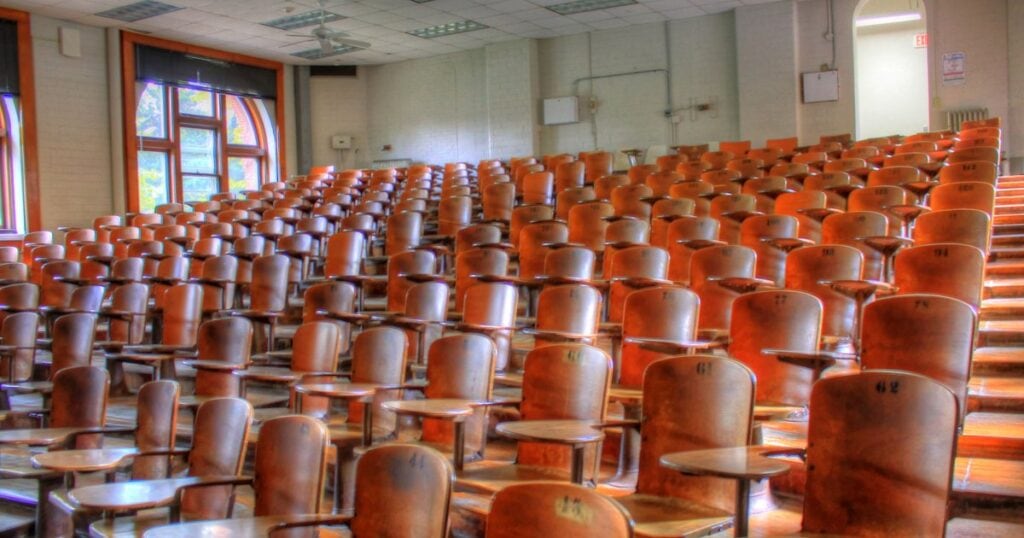 Άδειο πανεπιστημιακό αμφιθέατρο με ξύλινα καθίσματα σε σειρές και φως από τα παράθυρα.