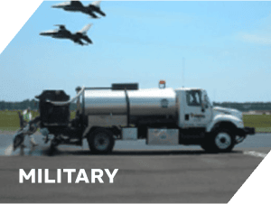 Military aircraft flying over a ground support vehicle at an airfield.