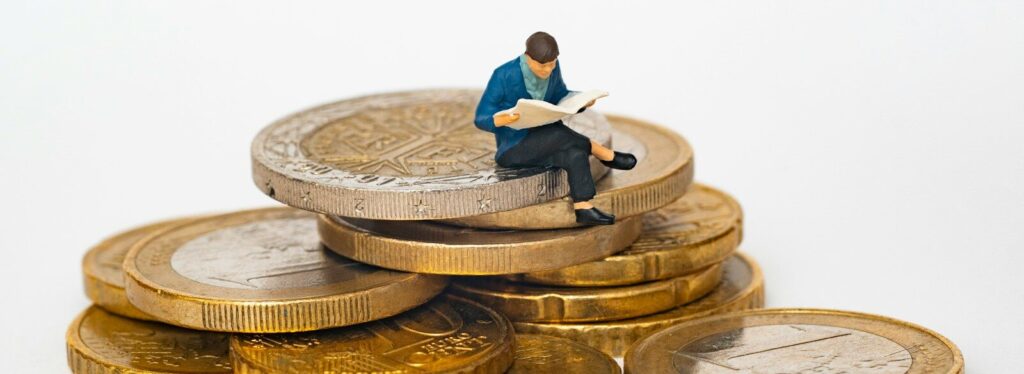 Miniature person sitting on stack of coins reading newspaper
