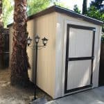 Compact outdoor storage shed with beige siding and black trim, situated in a backyard with palm tree.