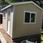 Compact beige backyard shed with white trim and a window, situated on a raised foundation in a garde.