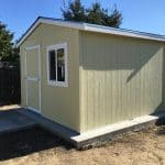 Garden shed with window and door, painted in light yellow, situated on a concrete slab in a backyard.