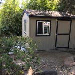 Small outdoor garden shed with gray siding, black trim, and windows in a backyard setting.