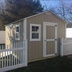 Garden shed with beige siding and white trim, surrounded by a white picket fence. Perfect for storag.