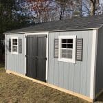 Garden shed with black double doors, white-framed windows with shutters, and gray siding, set in a b.