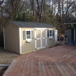 Garden shed with double doors, windows, and beige siding, situated on a wooden deck in a backyard.