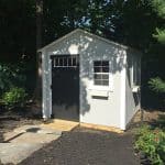 Garden shed with windows, black door, and white exterior in a lush backyard setting.