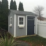 Garden shed with gray siding, white trim, and dark blue door, situated in a backyard with a white fe.