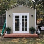 Garden shed with white double glass doors, surrounded by potted plants, situated on a wooden deck in.