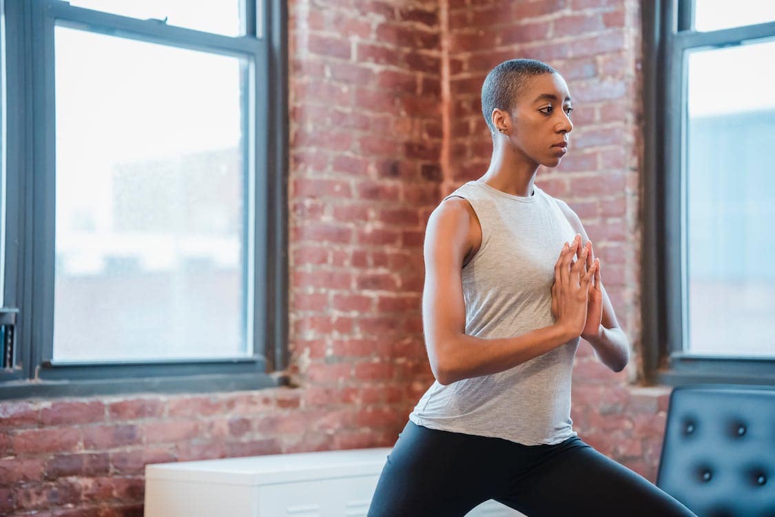 A woman with close-cropped hair practices yoga in a home automated gym, complete with large windows and rustic brick walls.