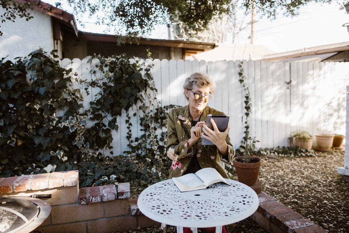An older woman sits on her bright patio and uses a tablet to check how smart home energy solutions have saved her money.