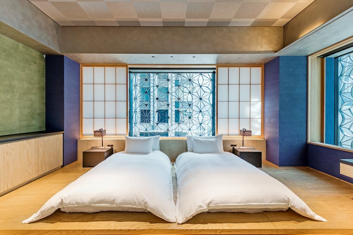 A bright, wide and colorful smart bedroom holds two mattresses, blue and purple curtains and elaborate windows.