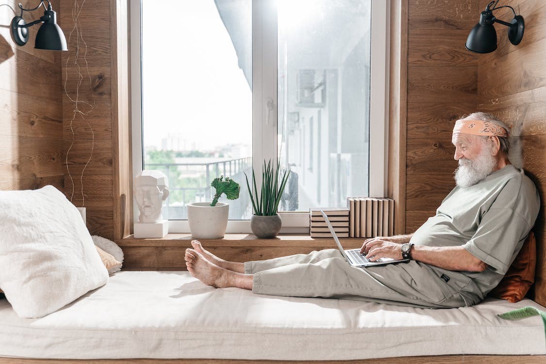 An older adult man checks his laptop while sitting on a comfortable bed with white sheets in front of a bright, wide window.